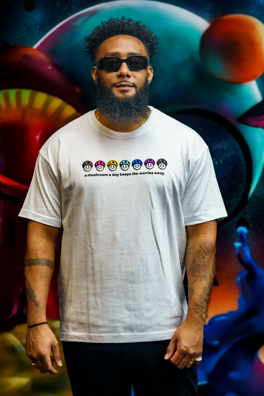 A Mushroom a Day Heavyweight Tee in white with graphic showing a row of colorful mushrooms worn by a man standing in front of a mural painting