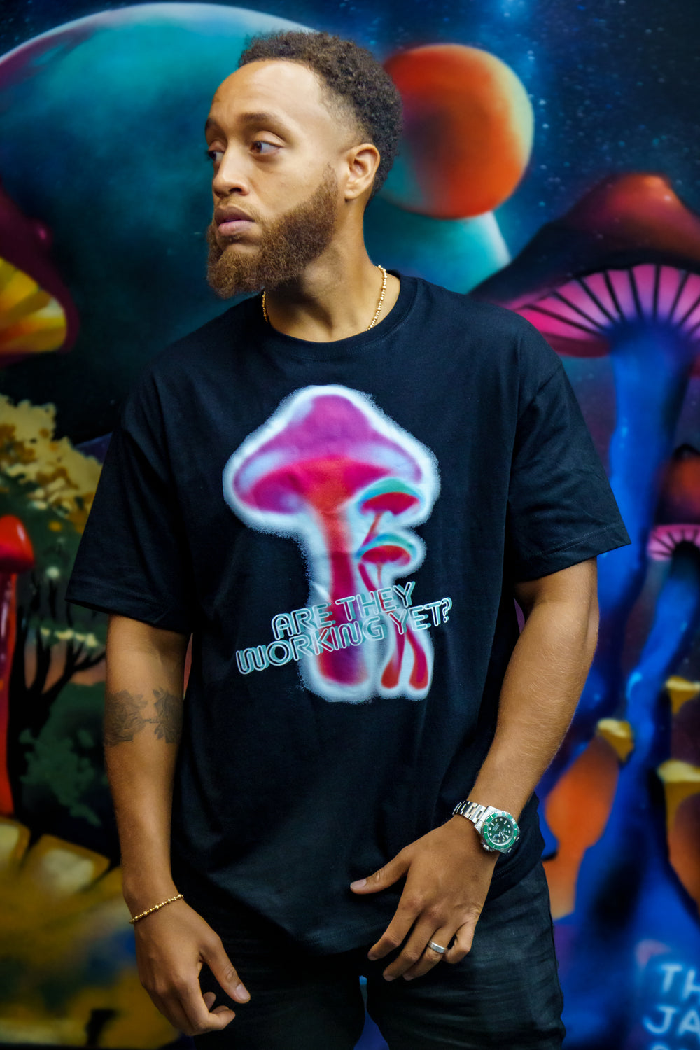 Are They Working Yet? Heavyweight Tee in black with a colorful mushroom graphic worn by a man standing in from of a mural