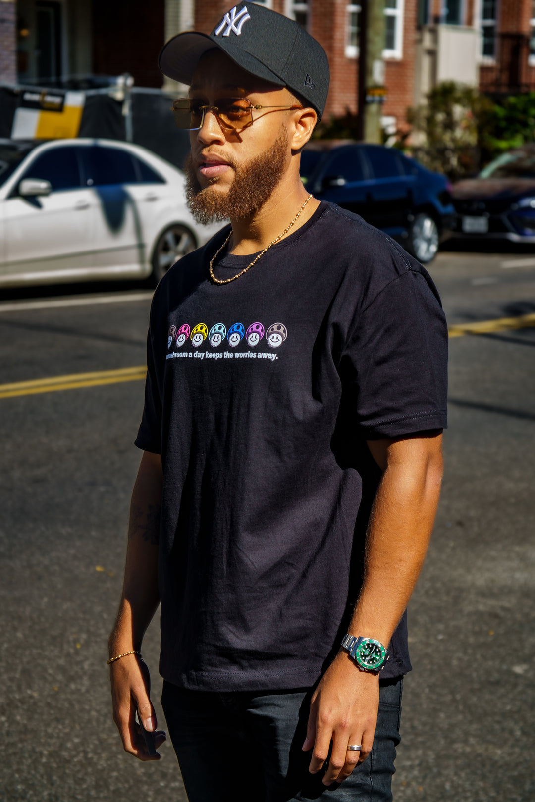 A Mushroom a Day Heavyweight Tee in black with graphic showing a row of colorful mushrooms worn by a man in a city